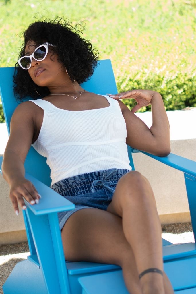 Black women laying on blue chair wearing a White Ribbed Body Suit and high waist shorts and white cat eye sunglasses